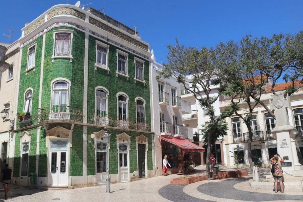 Una plaza en el casco antiguo de Lagos, Portugal, con casas encaladas, un edificio de tres pisos cubierto con azulejos verdes, algunos árboles en el medio de la plaza y una mujer que toma fotografías de una estatua