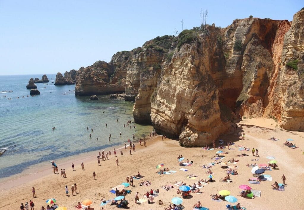 As águas cristalinas e as enormes falésias de calcário amarelo da Praia Dona Ana, Lagos, Portugal, com pessoas na água, caminhando na praia e tomando banho de sol em toalhas ou debaixo de guarda-sóis de cores diferentes