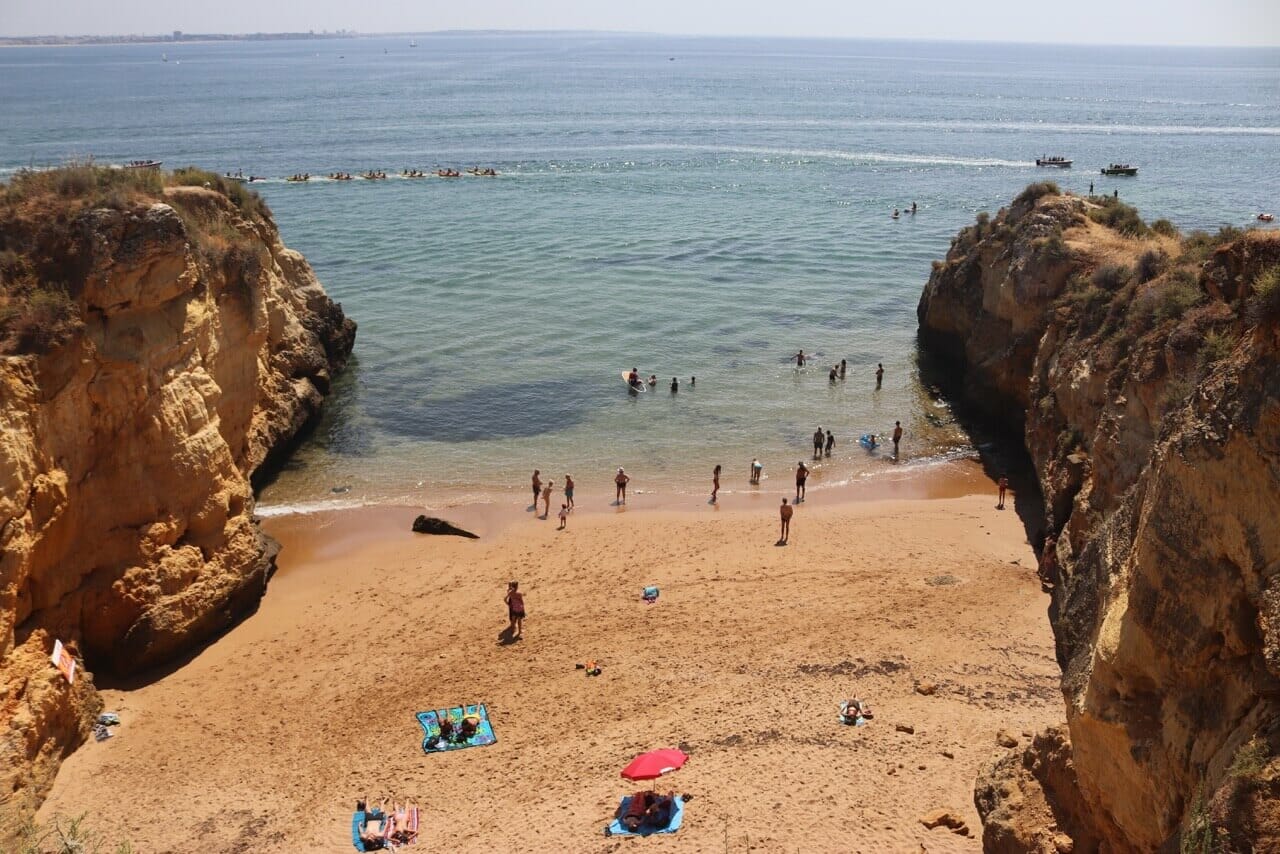 parte da Praia dos Estudantes, Lagos, com uma praia rodeada por falésias, pessoas a apanhar banhos de sol na areia ocre e outras na água