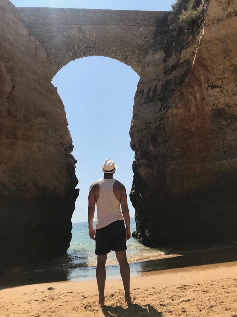 un hombre vestido con pantalones cortos azules, una camiseta sin mangas blanca y un sombrero blanco frente a un arco en la Playa de los Estudiantes, Lagos, Portugal