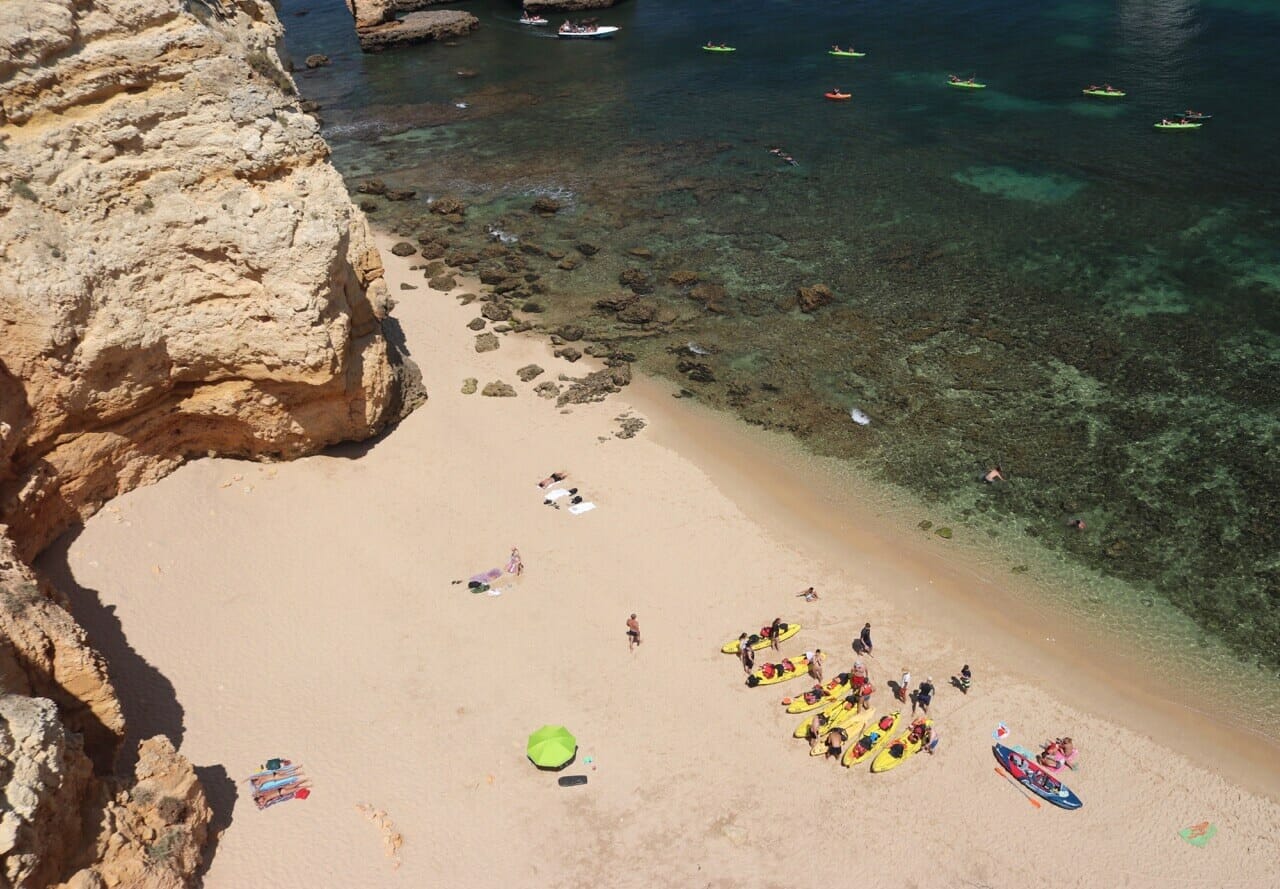 Aerial view of Praia Grande with a beige-orangeish cliff, people laying down on towels on the sand near yellow kayaks, the crystalline water with some coral reefs and some green kayaks sailing on the water