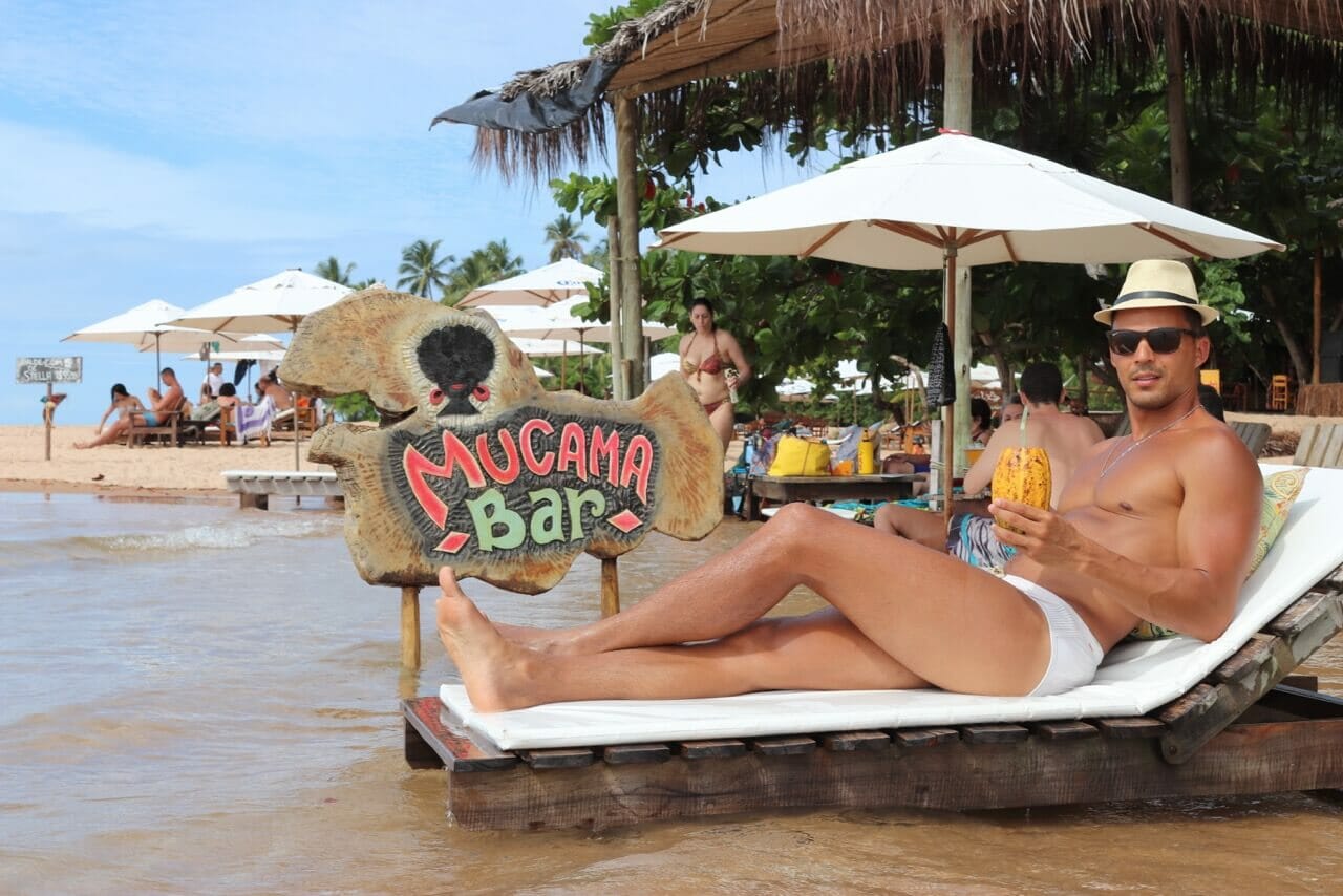 Pericles Rosa usando óculos escuro, chapéu e uma sunga branca com uma drink no cacau sentando em uma espreguiçadeira na Ponta do Mutá, em Barra Grande, Bahia