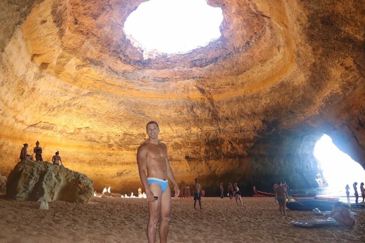 Some people walking and posing for pictures inside Benagil Cave, Portugal, which has golden-coloured walls with a round hole in the ceiling and an archway