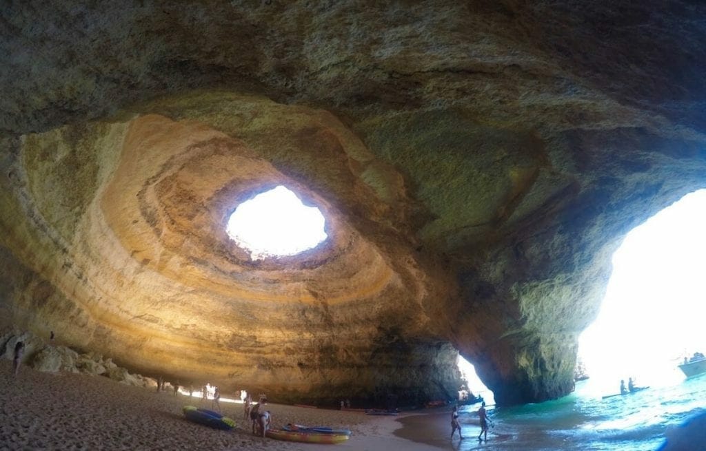 Some people and kayaks inside Benagil, a sea cave in Portugal that has golden walls, round ceiling with a hole in the middle and two archways