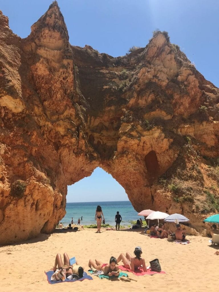 Tres mujeres tomando el sol, cuatro sombrillas y una chica caminando a través de un arco majestuoso en Praia dos Três Irmãos, Portimão, Portugal