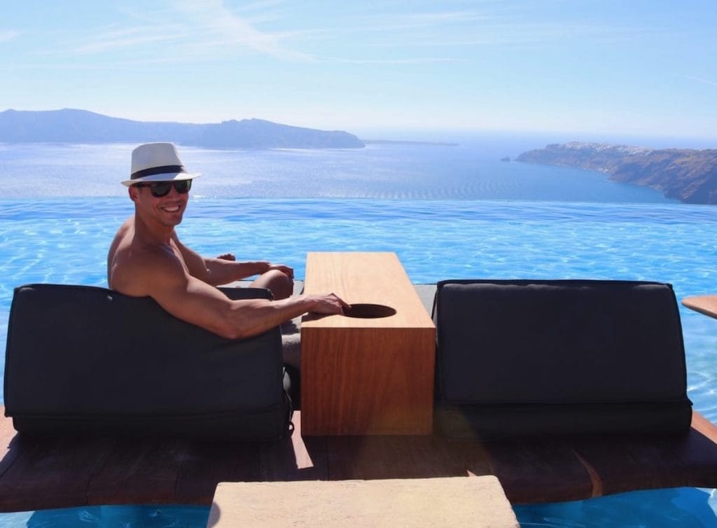 Pericles Rosa usando um chapéu branco e óculos de sol sentado em uma espreguiçadeira na piscina de borda infinita do hotel Cavo Tagoo, em Santorini, Grécia