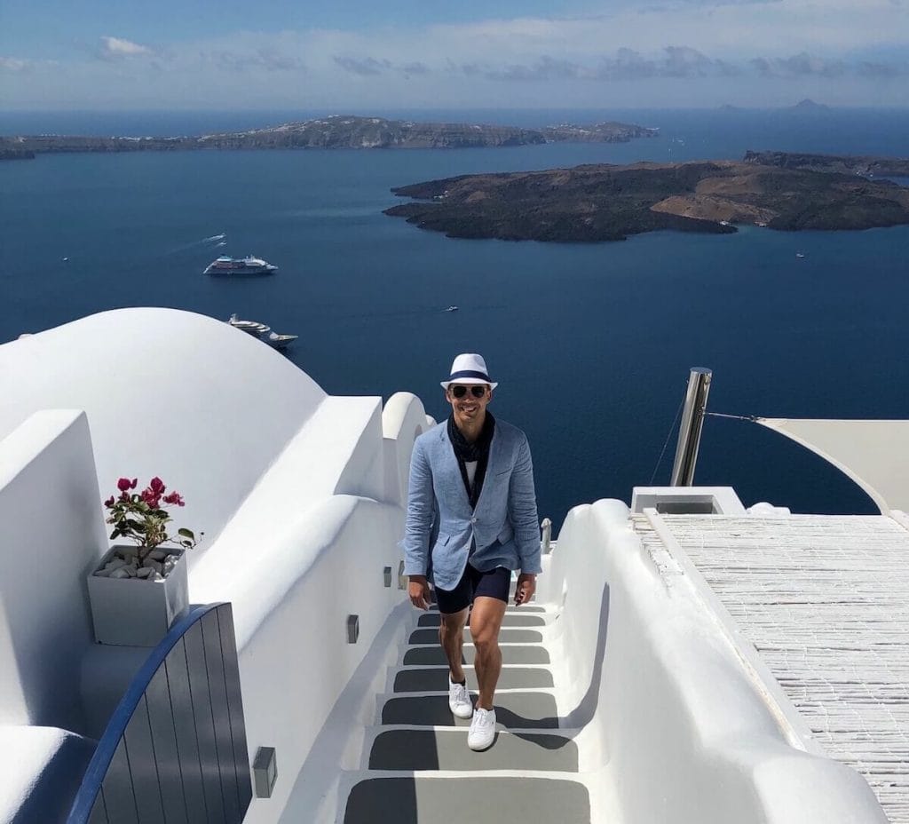 A man wearing a white hat, sunglasses, a light-blue blazer, a marine blue scarf, marine blue shorts and white trainers walking on the stairway of the Katikies Chromata Hotel, in Imerovigli, Santorini, with the Aegean Sea in the background 