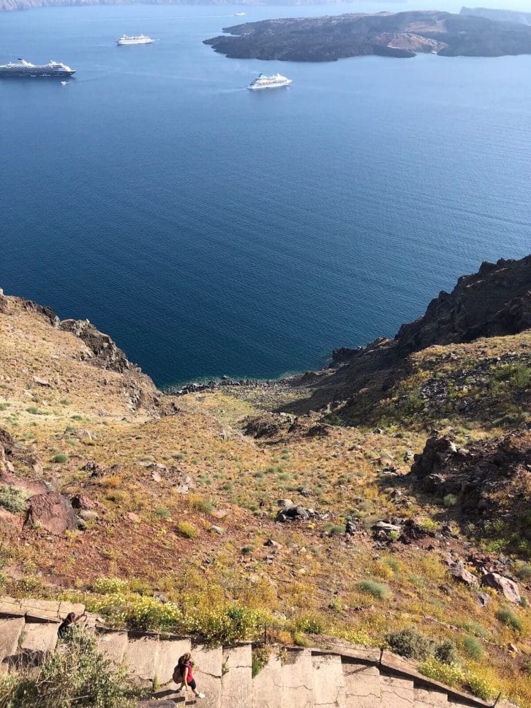 A woman walking down the stair to Skaros Rock, Imerovigli, Santorini, with the Nea Kameni Island and the Aegean Sea in the background