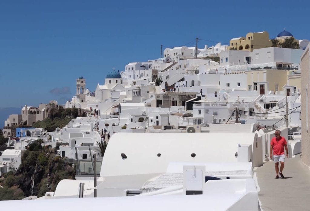 A man wearing a light red t-shirt and beige short walking on a path in the village of Imerovigli, Santorini, with several whitewashed houses and a blue-domed church surrounding him