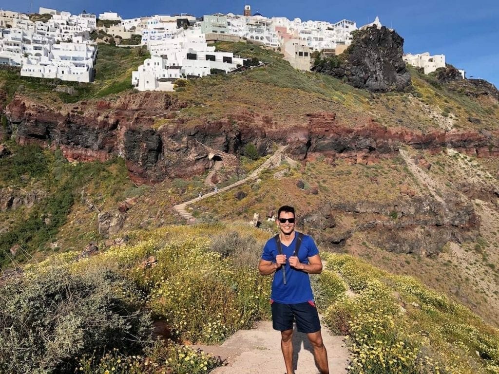 Pericles Rosa wearing a blue t-shirt, blue short and sunglasses hiking Skaros Rock and whitewashed houses of the village of Imerovigli, atop of a colossal red cliff covered low vegetation behind him