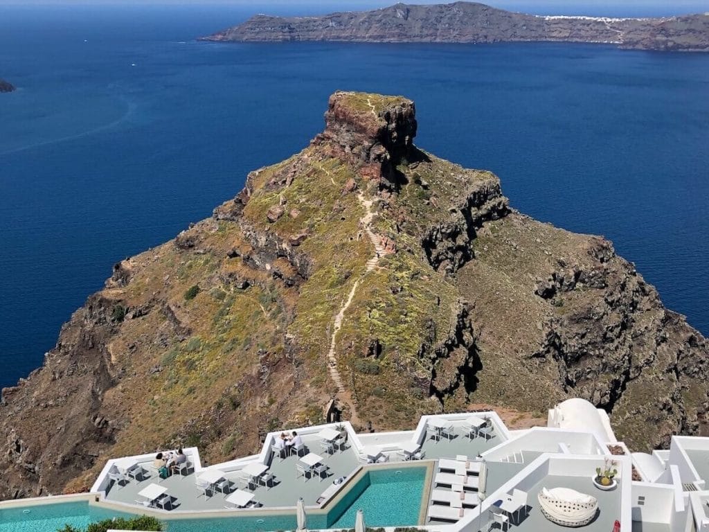 A swimming pool and a balcony with tables of a hotel in Imerovigli, Santorini, with the Skaros Rock and the Aegean Sea in the background