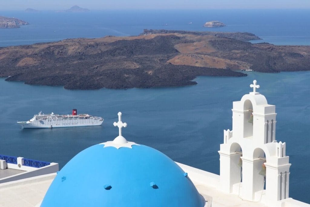 The blue-domed church of Agios Theodori in the village of Firostefani, Santorini, a cruise ship sailing in the Aegean Sea and the island of Nea Kameni in the background