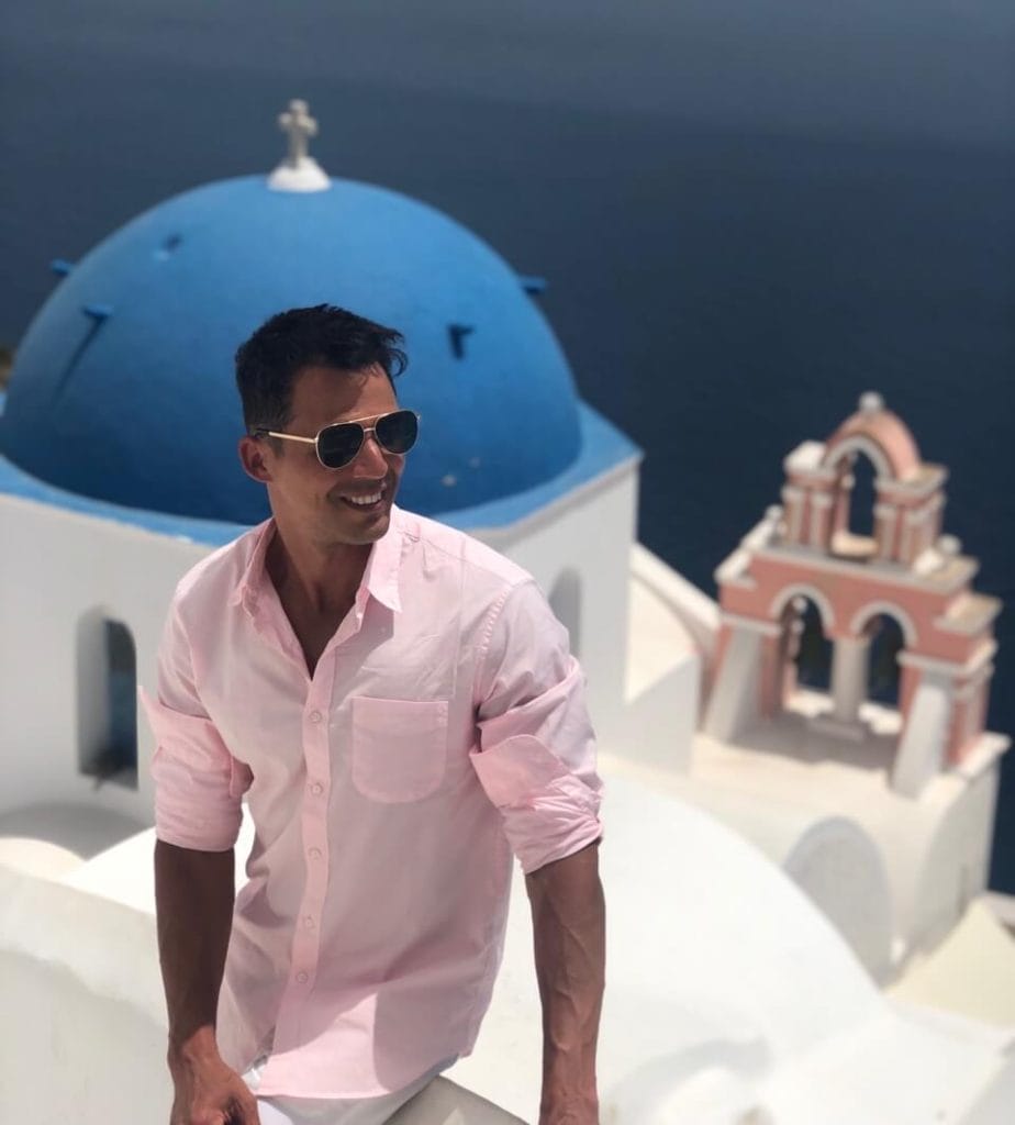 A man wearing sunglasses and a light pink shirt sitting on a white wall, a blue-domed church and a salmon bell tower behind him, in the village of Oia, Santorini