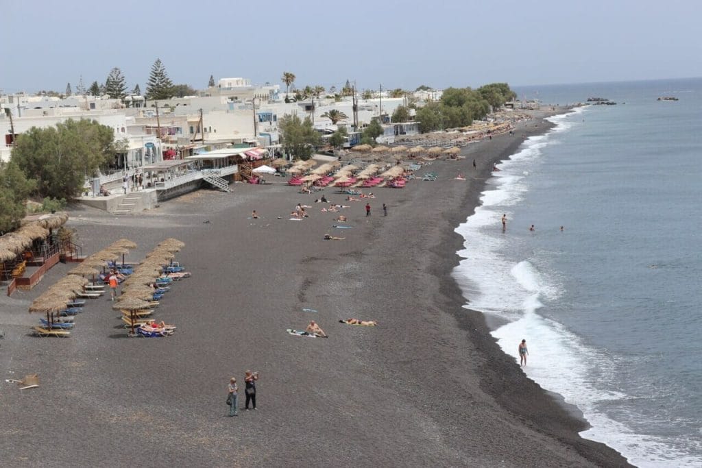 Algunas personas caminando en una playa de arena negra, sombrillas, tumbonas y casas encaladas en el pueblo de Kamari, Santorini, Grécia