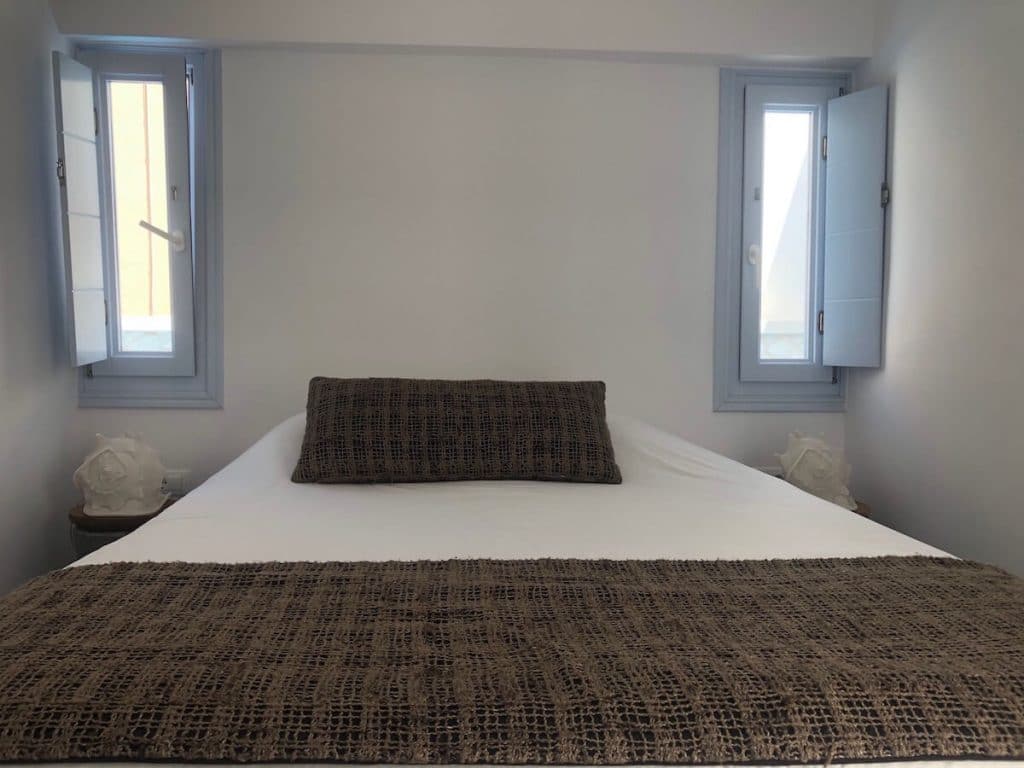 Una habitación del hotel San Giorgio en Fira, Santorini, con una cama doble con sábanas blancas y almohada marrón y pequeñas ventanas de color azul claro