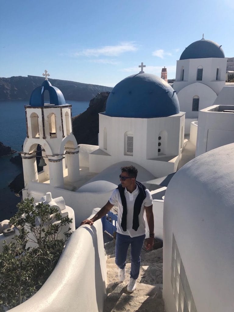 Um homem usando óculos escuros, polo branca, casaco azul marinho nos ombros e calça azul claro caminhando em um beco em Oia, Santorini, e uma igreja branca com três cúpulas azuis atrás dele