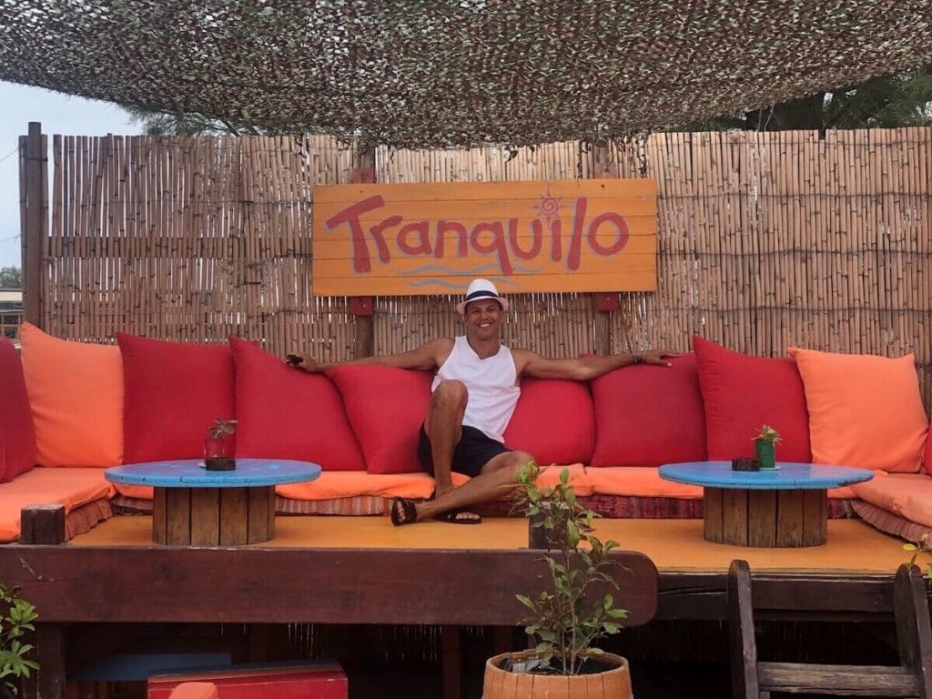 Péricles Rosa usando um chapéu branco, regata branca e shorts azuis sentado em um sofá laranja com almofadas vermelhas no Beach Bar Tranquilo em Santorini