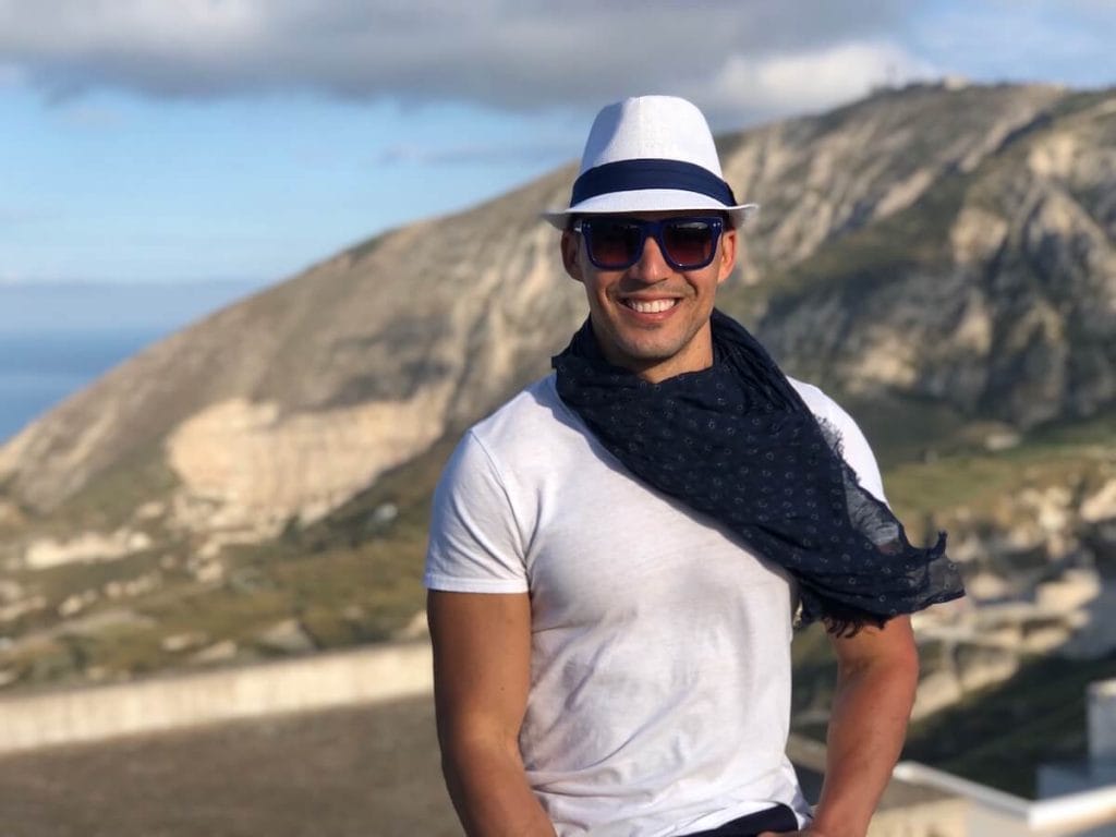 Un hombre de sombrero y camisa blanca y gafas de sol azul en la vila de Pyrgos, Santorini, con una montaña al fondo