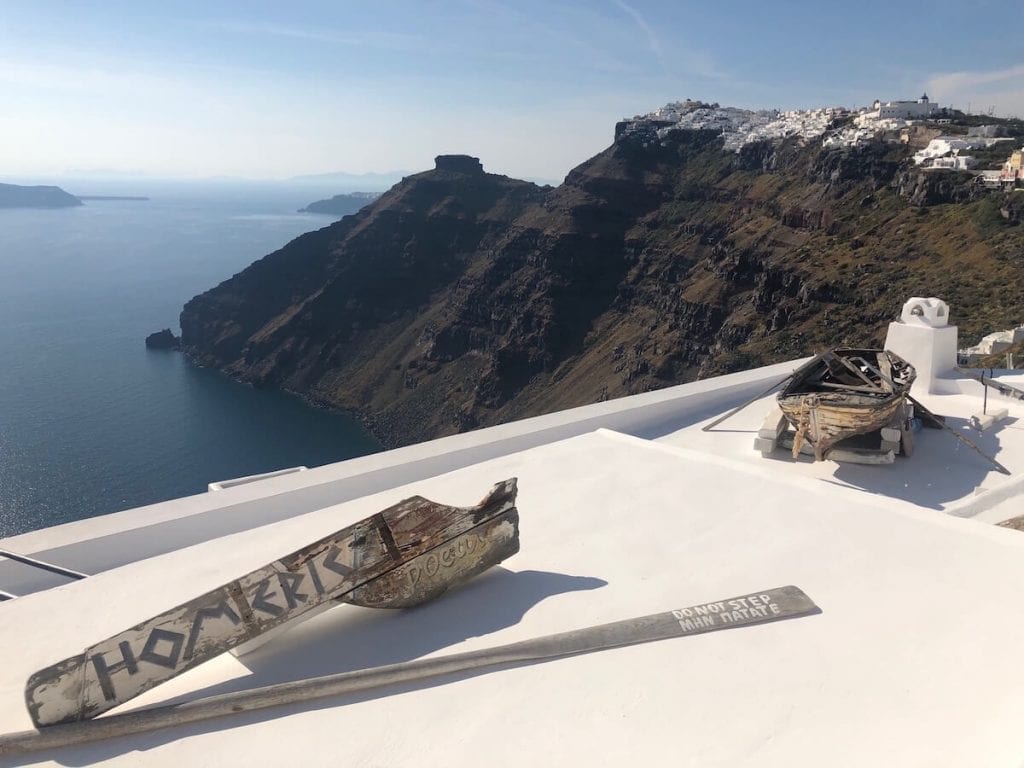 An old wooden boat on the rooftop of  Homeric Hotel Santorini, and the whitewashed houses of the villages of Firostefani and Imerovigli and the Agean Sea in the background