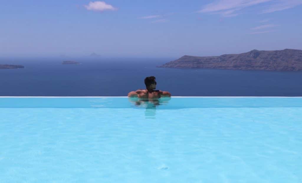 Pericles Rosa standing inside an infinity swimming pool at CSky Luxury Hotel in Santorini, with the Aegean Sea in the background 