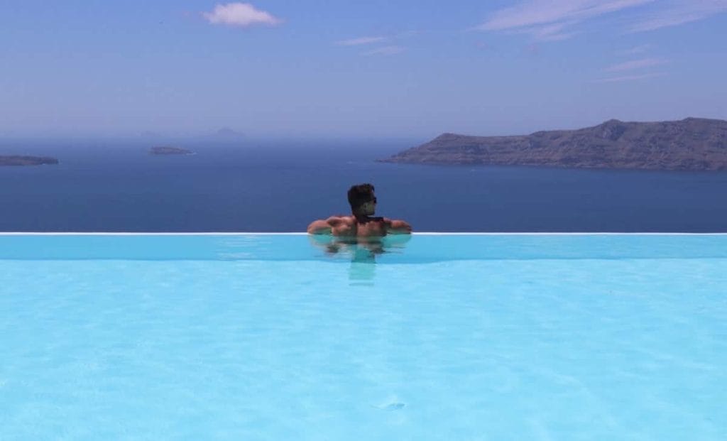 Pericles Rosa standing inside an infinity swimming pool at CSky Luxury Hotel in Santorini, with the Aegean Sea in the background