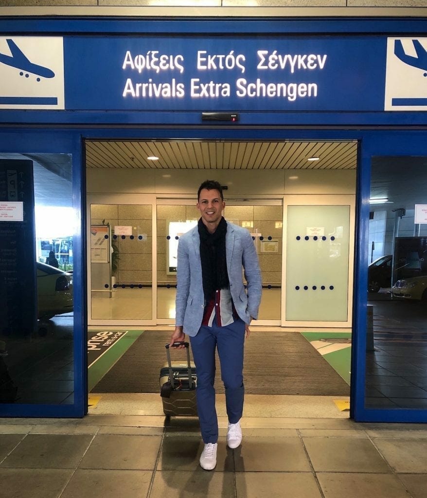 Pericles Rosa usando um blazer azul claro, cachecol azul escuro, camisa listrada, calça azul clara e tênis branco puxando uma mala no aeroporto internacional de Atenas, Grécia