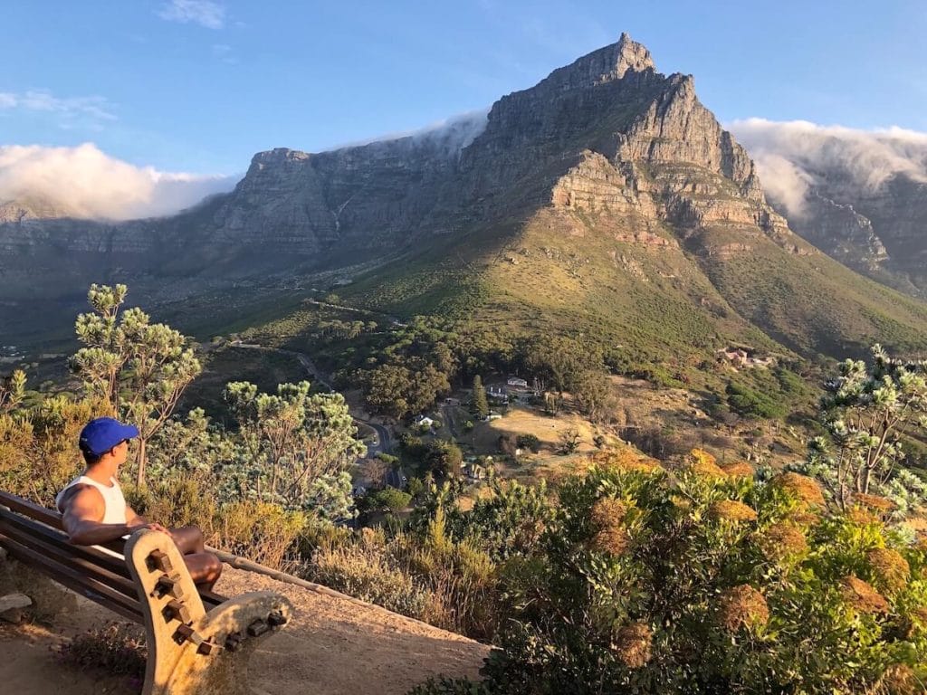 a man wearing a white tank top and blue hat seating on beach on Lions head trail admiring the Table Mountain and one of the best views in Cape Town