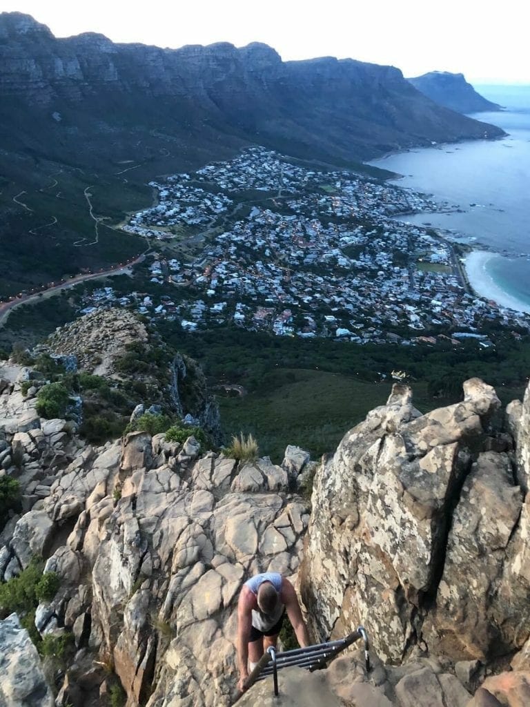a man climbing the last ladder of the Lions Head hike alone and Camps Bay Beach and the Twelve Apostles on the background