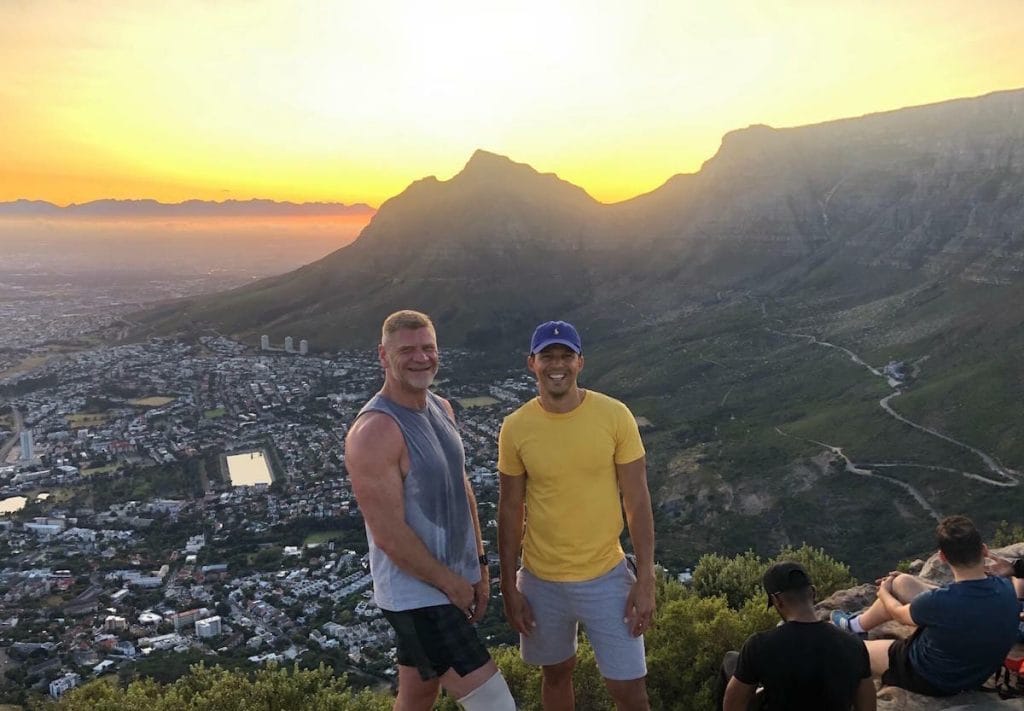 two men standing up and smiling at the Lion's Head summit while the sun is rising behind them and the Table Mountain