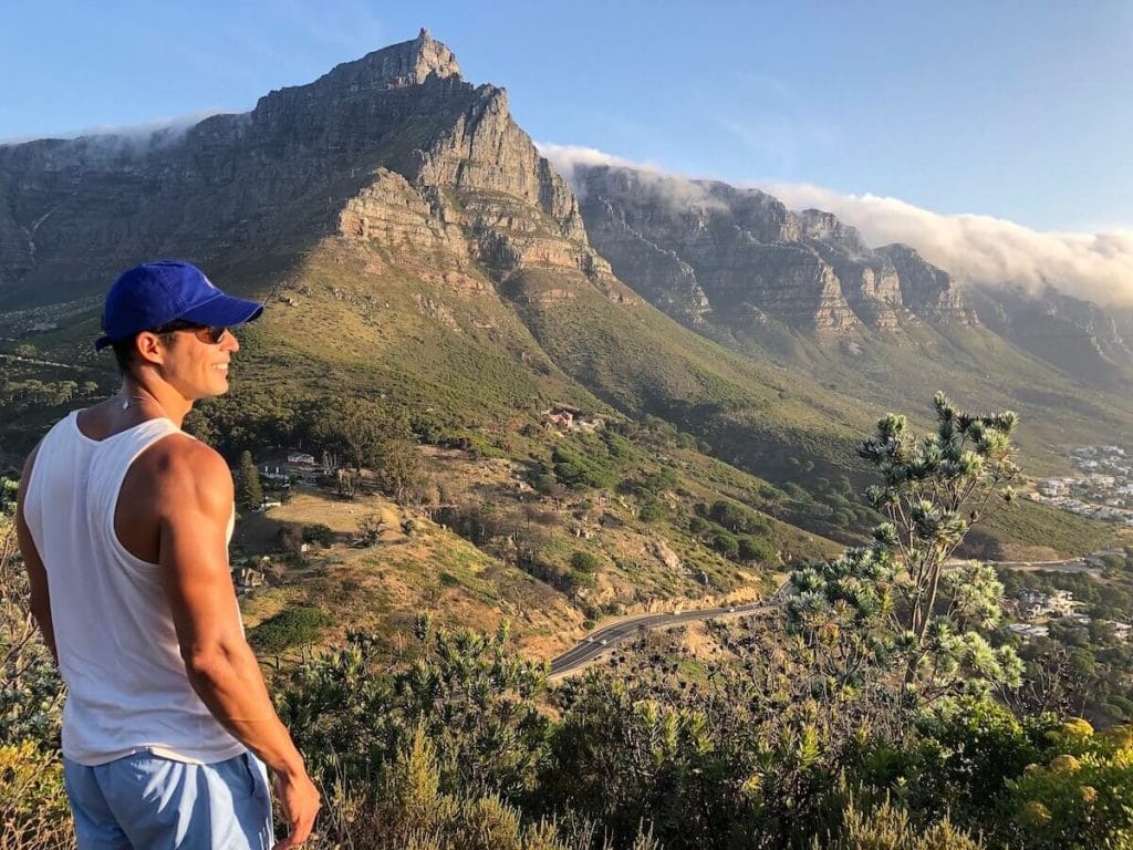 Pericles Rosa wearing a light blue short, a white tank top and a marine blue hat admiring the view of the Table Mountain and Twelve Apostles from Lion's Head Hiking Trail