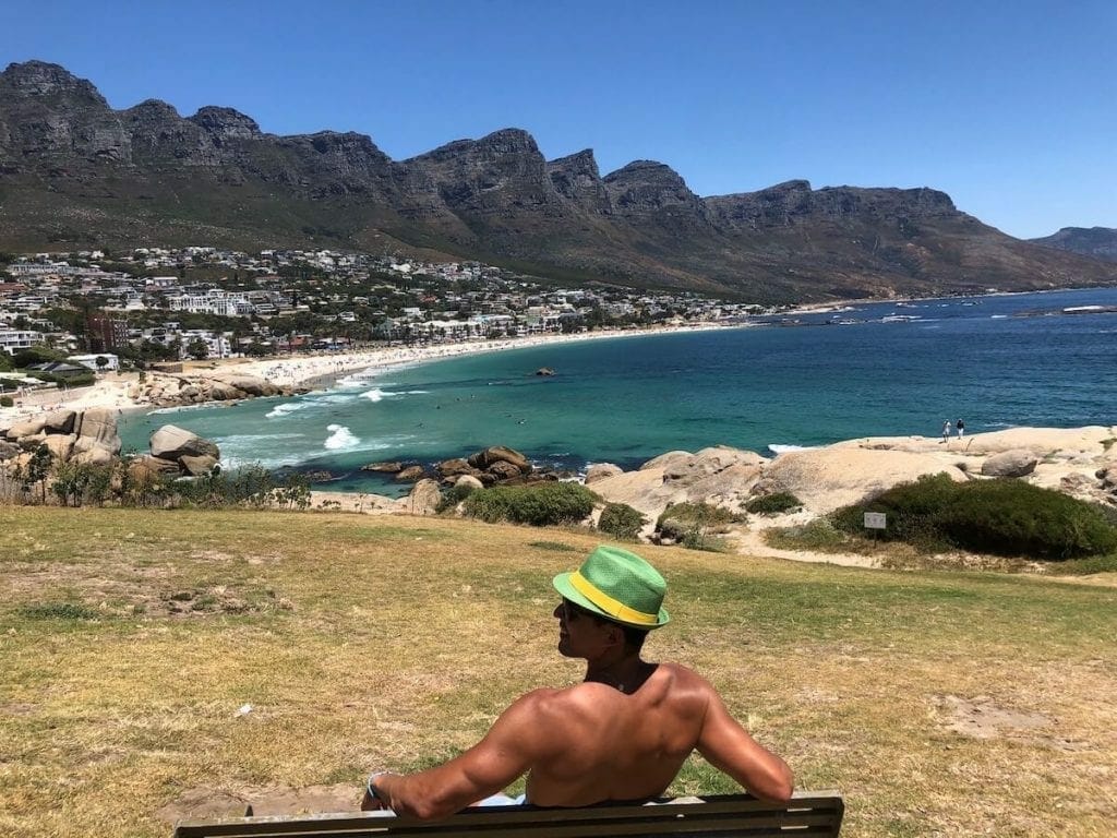 Pericles Rosa wearing a green and yellow hat sitting on a bench in front of Camps Bay Beach in Cape Town, a beach that has white soft sand, crystal clear blue water and is backed up by the Twelve Apostles Mountain