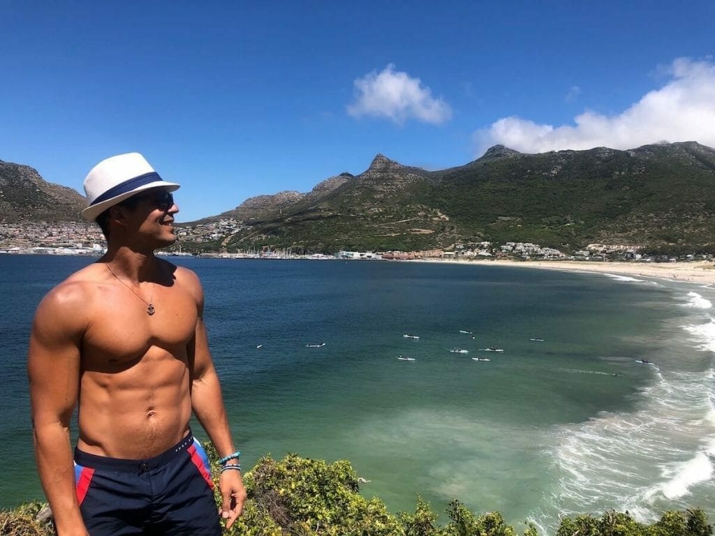 Pericles Rosa wearing a  white hat, sunglasses and blue shorts admiring the view of the top of a hill at Hout Bay Beach, Cape Town, South Africa