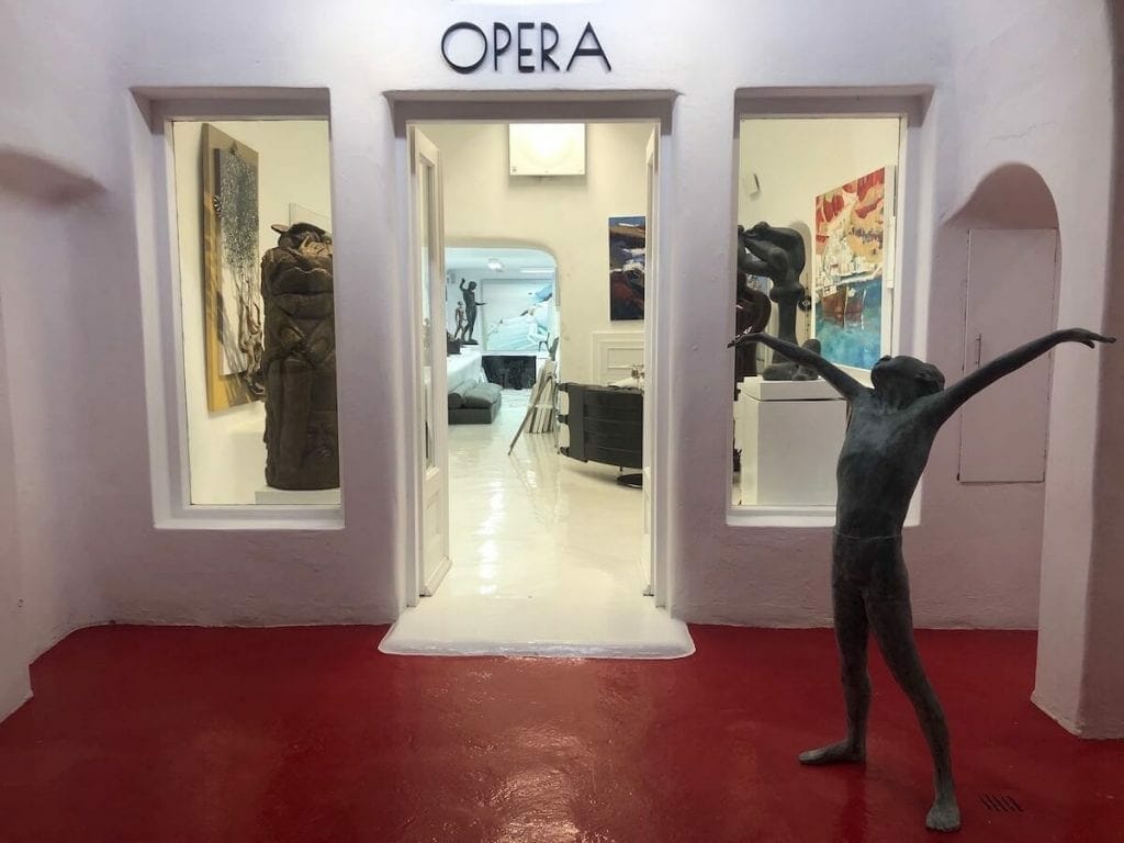 A statue at the entrance of Opera Art Gallery in Oia, Santorini, and other several artworks inside the gallery in the background
