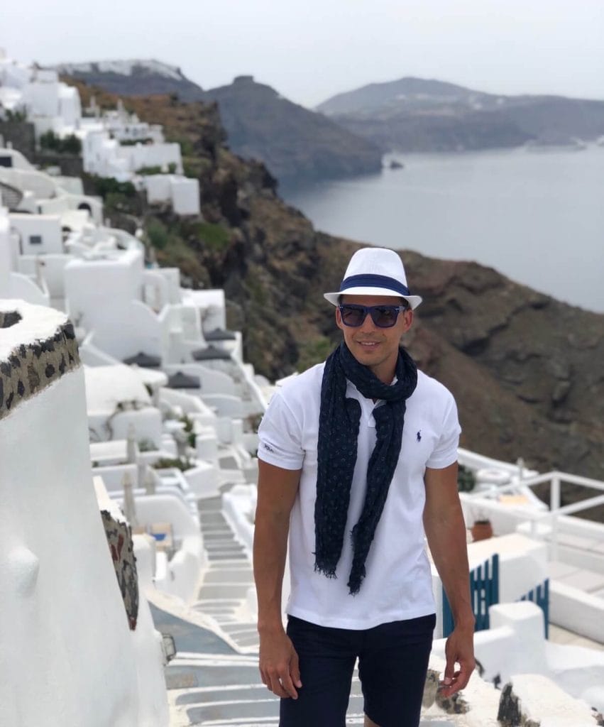 Um homem usando um chapéu branco, óculos escuros, cachecol azul marinho, polo branca e short azul marinho em um beco em Oia, Santorini, com algumas casas caiadas de branco ao fundo