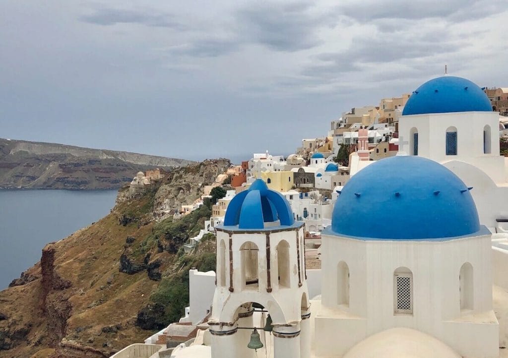 Una iglesia blanca con tres cúpulas azules e el pueblo de Oia al fundo, Santorini, Grecia