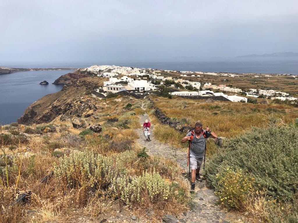 Dos personas con bastones caminando por el sendero entre Fira y Oia y el pueblo de Oia en el fondo