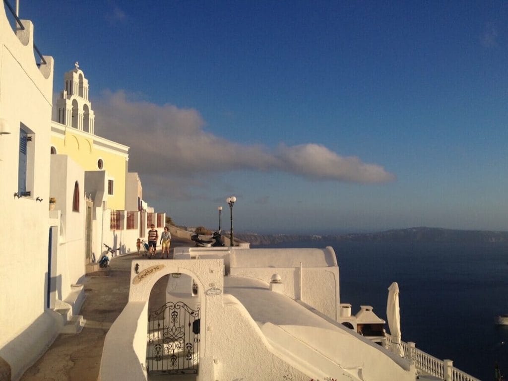 A couple walking by some whitewashed houses and an yellow church on the hiking trail from Fira to Oia and the Agean Sea in the background