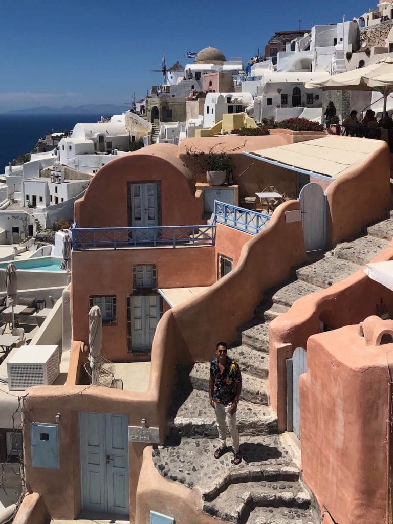 Un hombre de pie en las escaleras de un hotel con paredes de color salmón y algunas casas encaladas en el fondo, Oia, Santorini