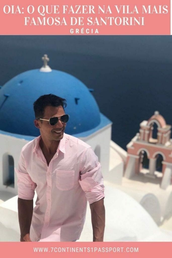 um homem usando óculos escuros e uma camisa rosa e uma igreja com cúpula azul com um campanário salmão e o Mar Egeu ao fundo, na cidade de Oia, Santorini