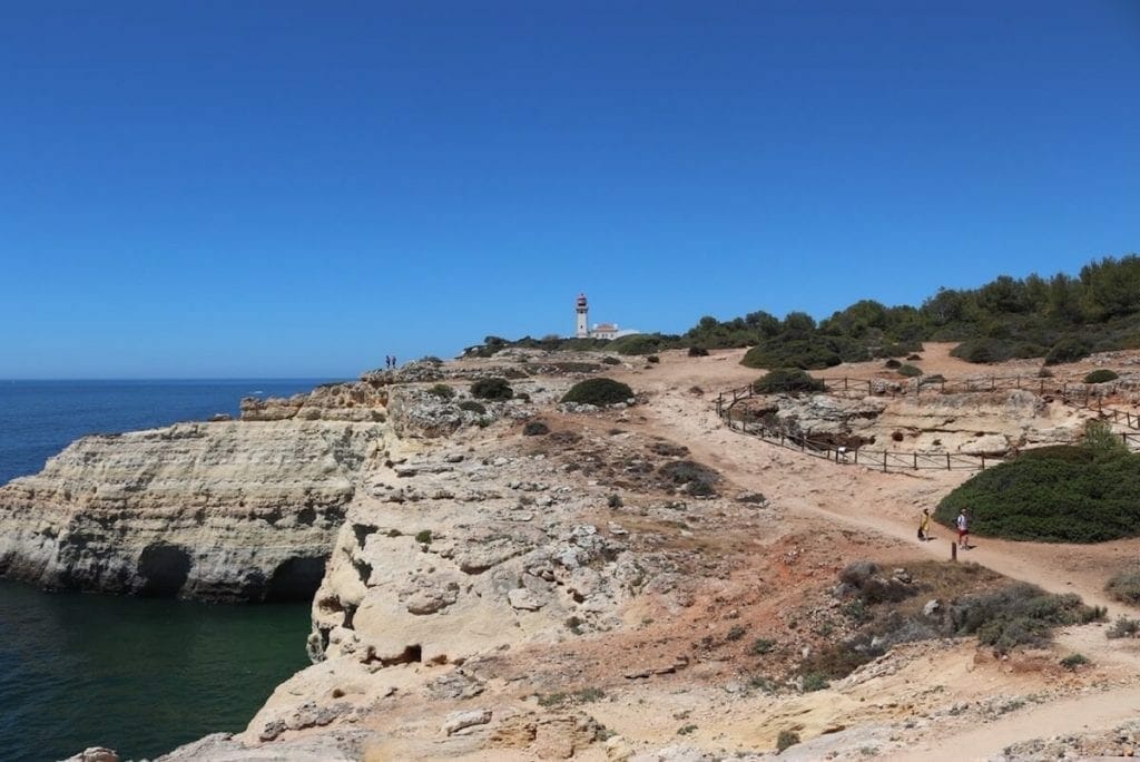 A man walking on the Seven Hanging Valleys trail, Portugal, that's an ocean path with some parts covered with low vegetation and has a lighthouse  