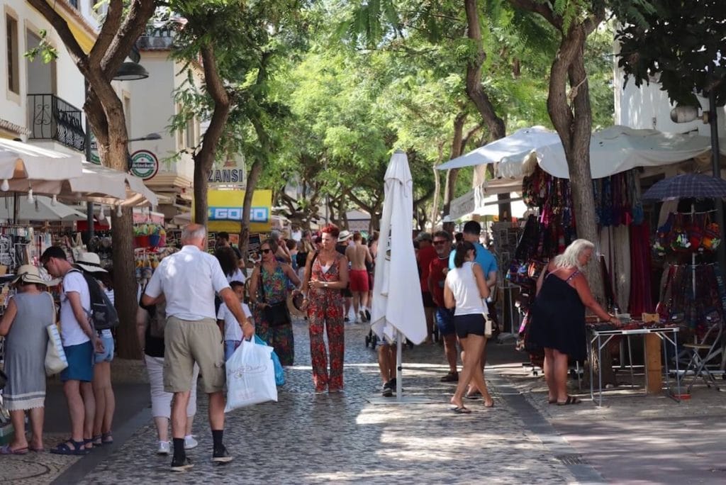 Pessoas fazendo compras numa rua movimentada da Cidade Velha de Albufeira com algumas lojas em ambos os lados e árvores