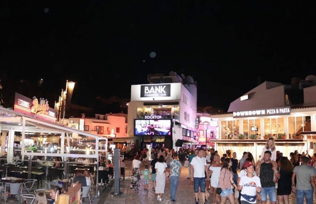 People walking in the evening on a square in the Old Town of Albufeira with some bars and restaurants with outdoor seating and a three-story bar with a rooftop in the middle of two streets in the background