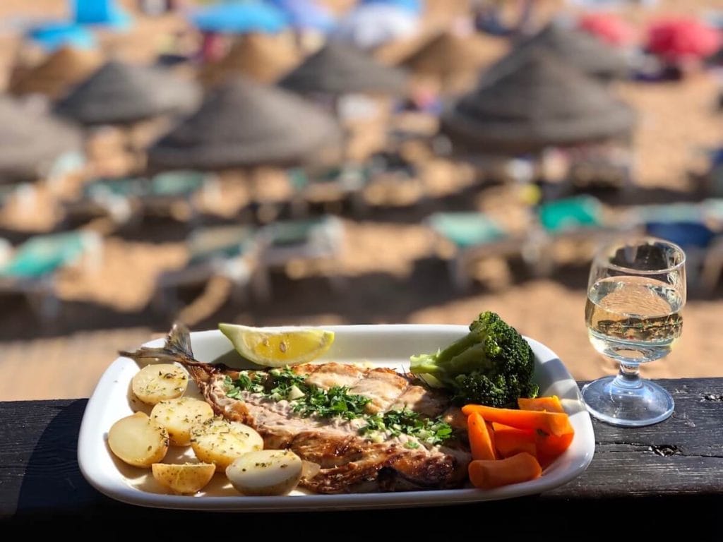 Um prato branco com peixes, batatas, brócolos, cenouras e limão, e um copo de vinho branco, com guarda-sóis e cadeiras da Praia da Coelha ao fundo