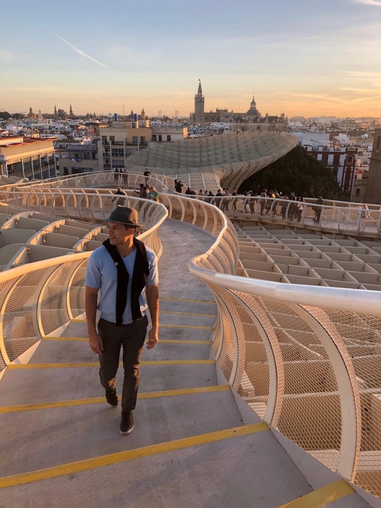 Pericles Rosa wearing a grey pants, light blue t-shirt and a grey hat walking on the metropol parasol and the city of seville in the background