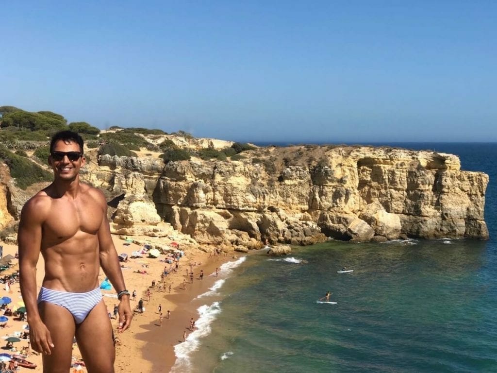 Pericles Rosa wearing a speedo and sunglasses of the to of a cliff on Praia da Coelha in Albufeira and in the background massive yellow limestone cliffs that surrounds the beach and people walking to beach and in the water