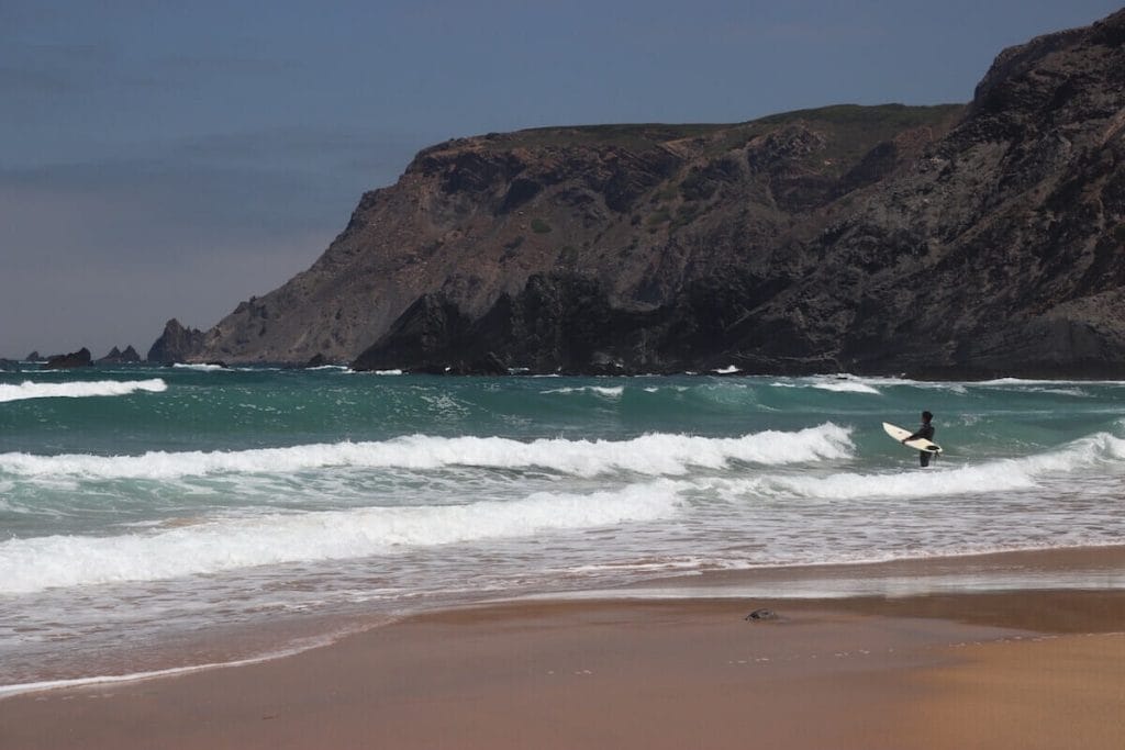 A man with a surfboard getting into the water on a beach in Sagres, Portugal