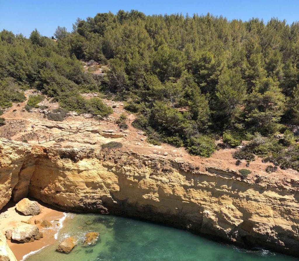 Part of the Seven Hanging Valleys Trail, Algarve, with a small beach with crystal-clear water surrounded by orange limestone cliffs some local vegetation covering the cliffs