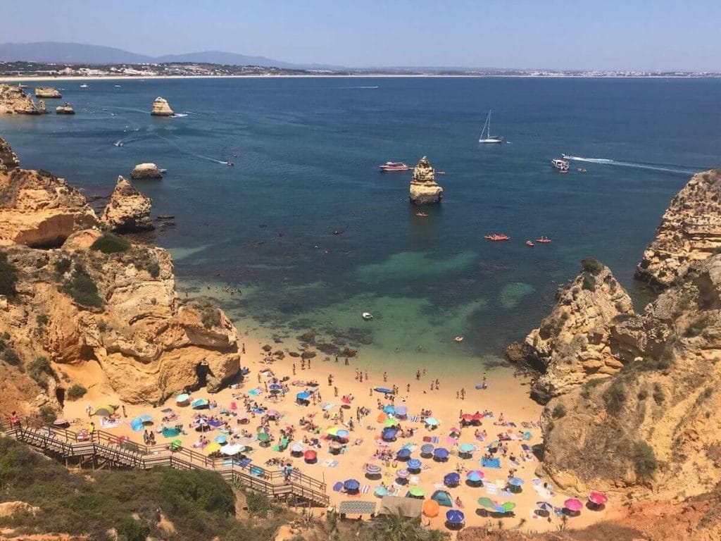 Playa Camilo, Lagos, Portugal, con varias sombrillas de diferentes colores, gente paseando por la playa que está rodeada por enormes acantilados de piedra caliza de color amarillo anaranjado y una escalera de madera que da acceso a la playa