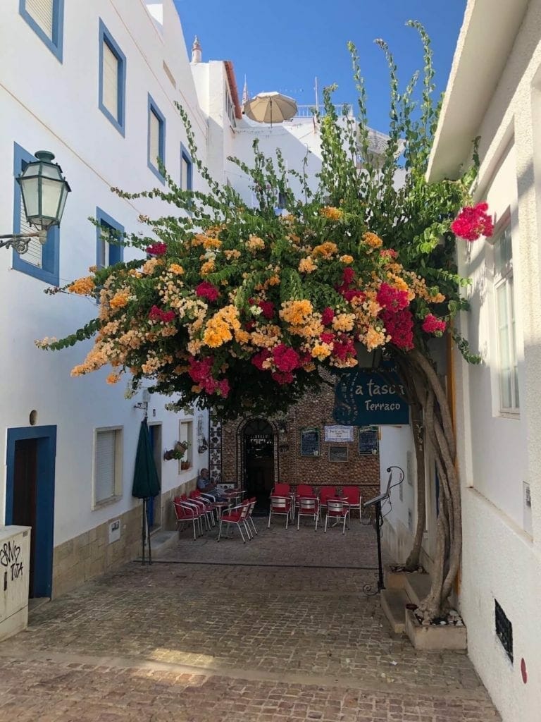 a cobblestone alley in Old Town Albufeira with a bougainvillea tree full of orange and red flowers, and a restaurant with outdoor seating in the background