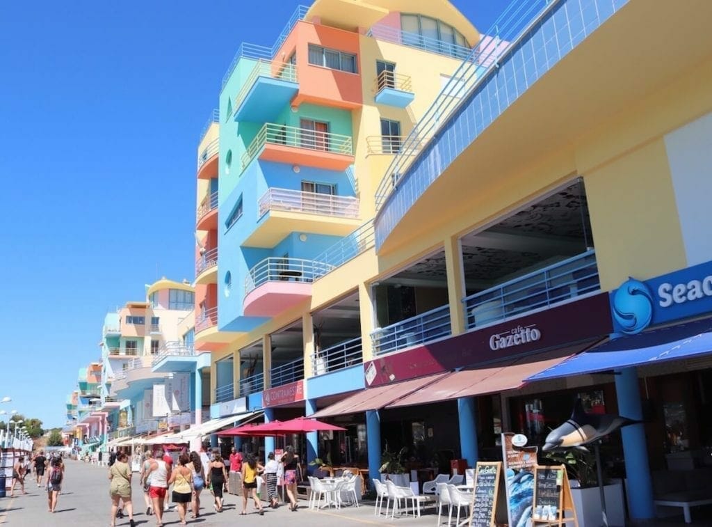 The Marina de Albufeira with its bars, restaurants with outside seatings, seven-storey colourful buildings, and some people walking